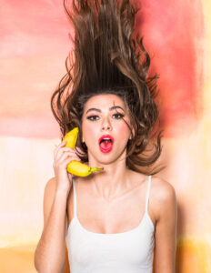 woman in a white tank top flips her hair while making a surprised face and holding a banana like a phone.