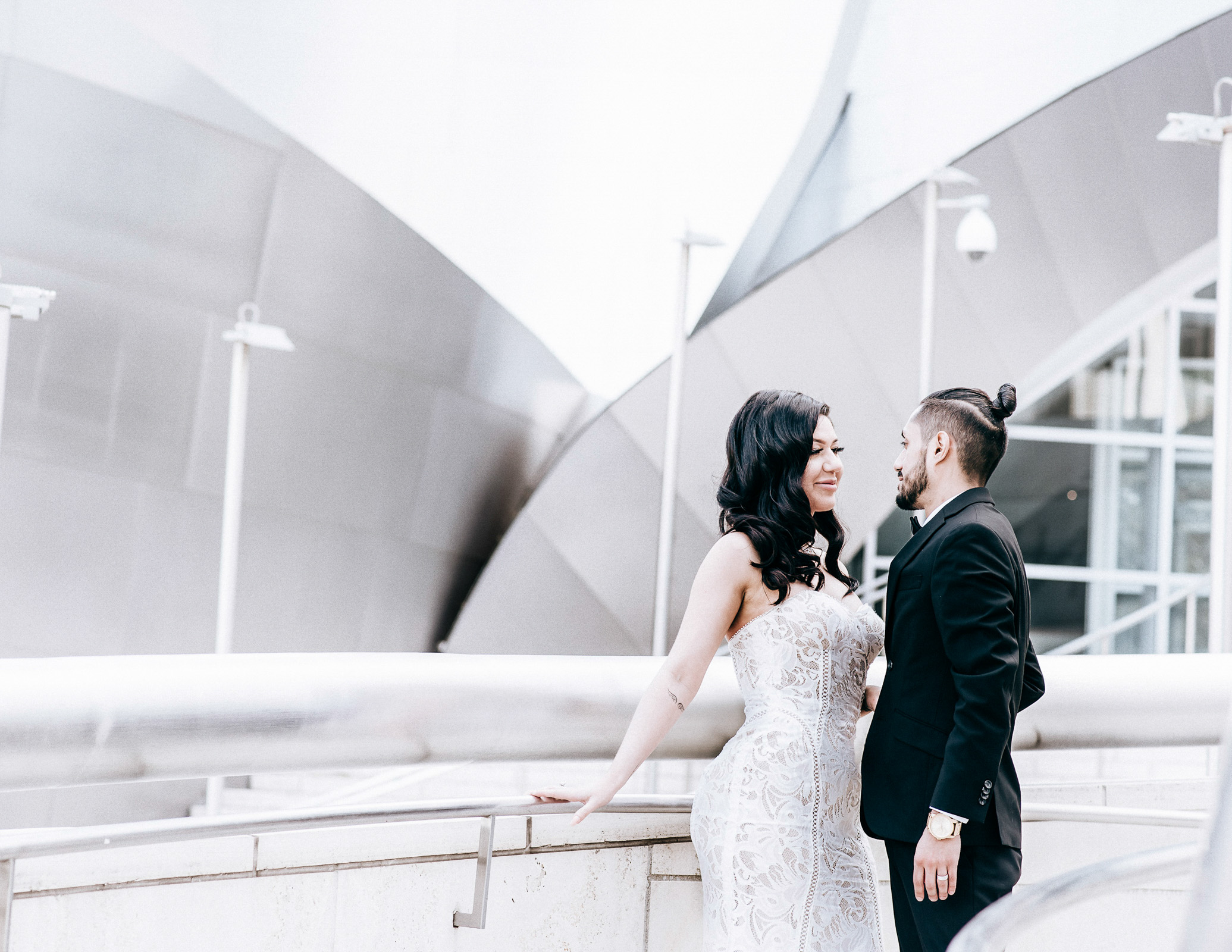 couple in formal wear standing by a railing, with a modern geometric building in the background.