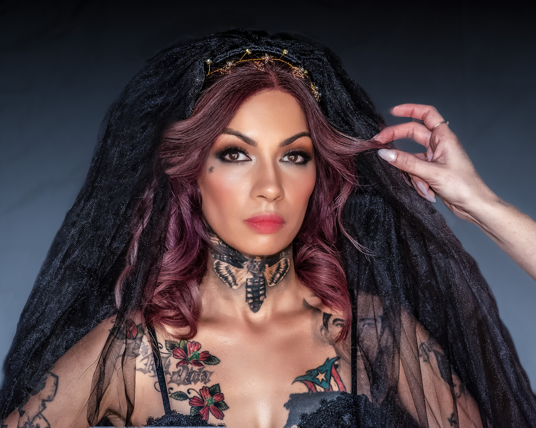 portrait of a tattooed woman with burgundy hair and a black lace veil, a hand touching the veil to frame her face.