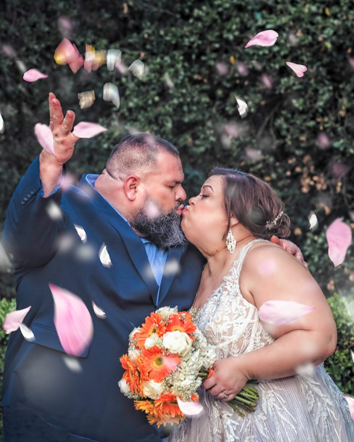 Bride and groom kiss outdoors under falling pink petals, bouquet of orange and white flowers in view behind them.