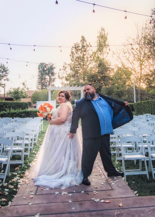 Bride in a white wedding gown holding an orange bouquet, posing with a man in a blue shirt and suit outdoors under string lights.