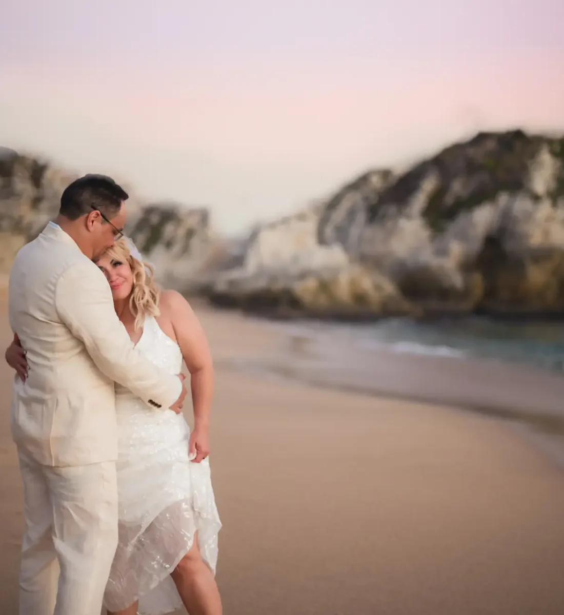 A couple in wedding attire embraces on a sandy beach at sunset, with rocky cliffs in the background.