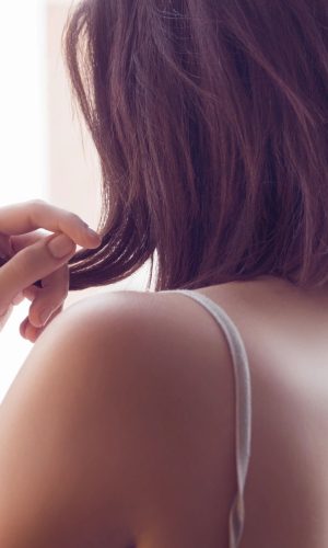 Back view of a person with shoulder-length brown hair wearing a light strap top, touching hair at the nape of the neck, seen from behind.